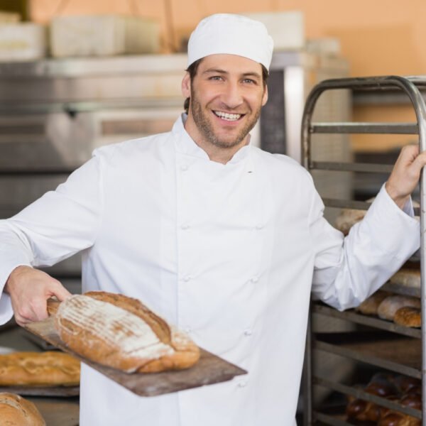 Un panadero sonriente sostiene una tabla con pan artesanal en un entorno de cocina limpio y profesional.
